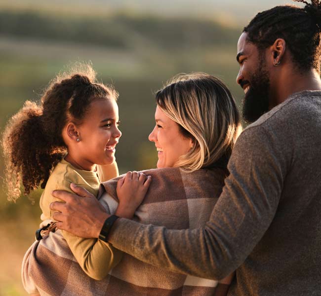Three people outdoors; one adult holding a child, another adult standing close